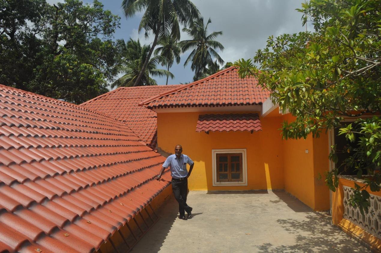 1 man standing by a bright orange building with red tiled roof surrounded by palm trees and greenery